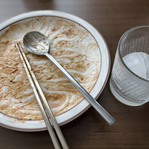 An empty plate with an empty cup, spoon, and chopsticks after eating all the fried noodles.