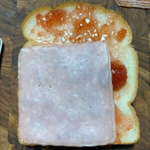 딸기잼을 바른 식빵 위에 슬라이스 햄 한 장을 올린 모습.