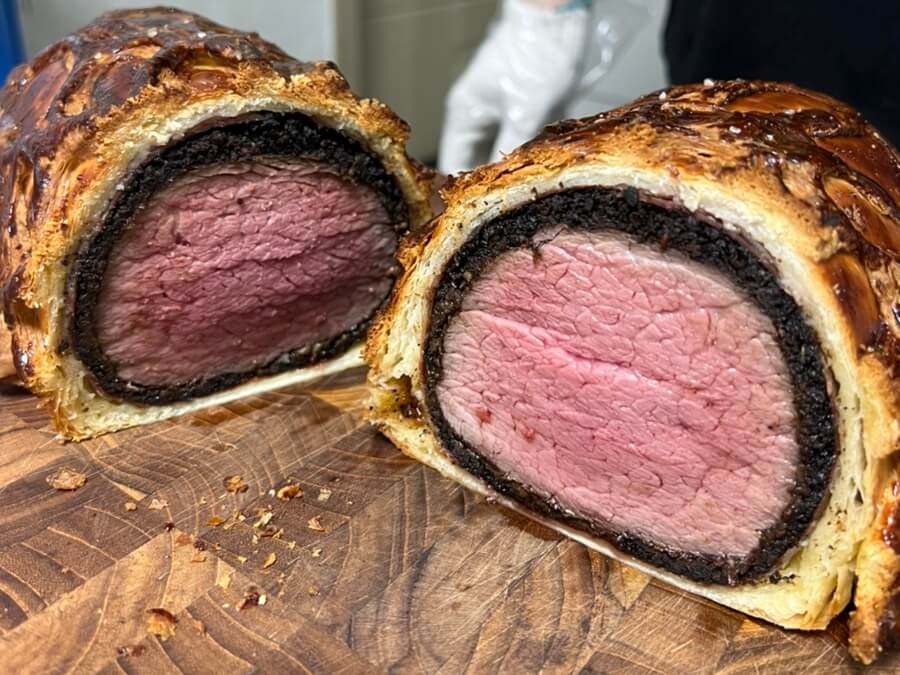 Receta de carne de buey Wellington al vacío. El buey Wellington terminado se corta por la mitad en una tabla de cortar marrón para mostrar la sección transversal.