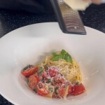 ブリーチーズパスタレシピを完成させた様子 白い皿の上に黄色いパスタの麺と赤いミニトマト、緑のバジルが乗せられ、その上に白いパルミジャーノ・レッジャーノチーズをすりおろしたものが乗っています。