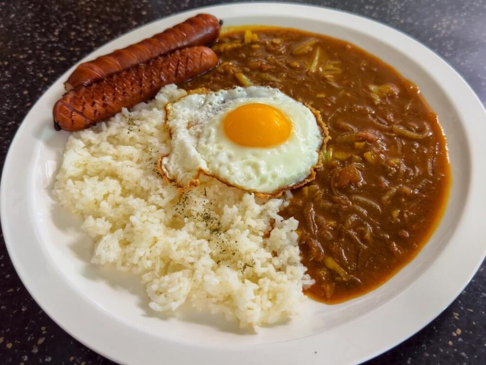 トマトカレーレシピを完成させた様子で、白い皿の上に白米ご飯と薄めのカレー、その横に焼きソーセージが2枚乗っている。真ん中には目玉焼きが乗っている。